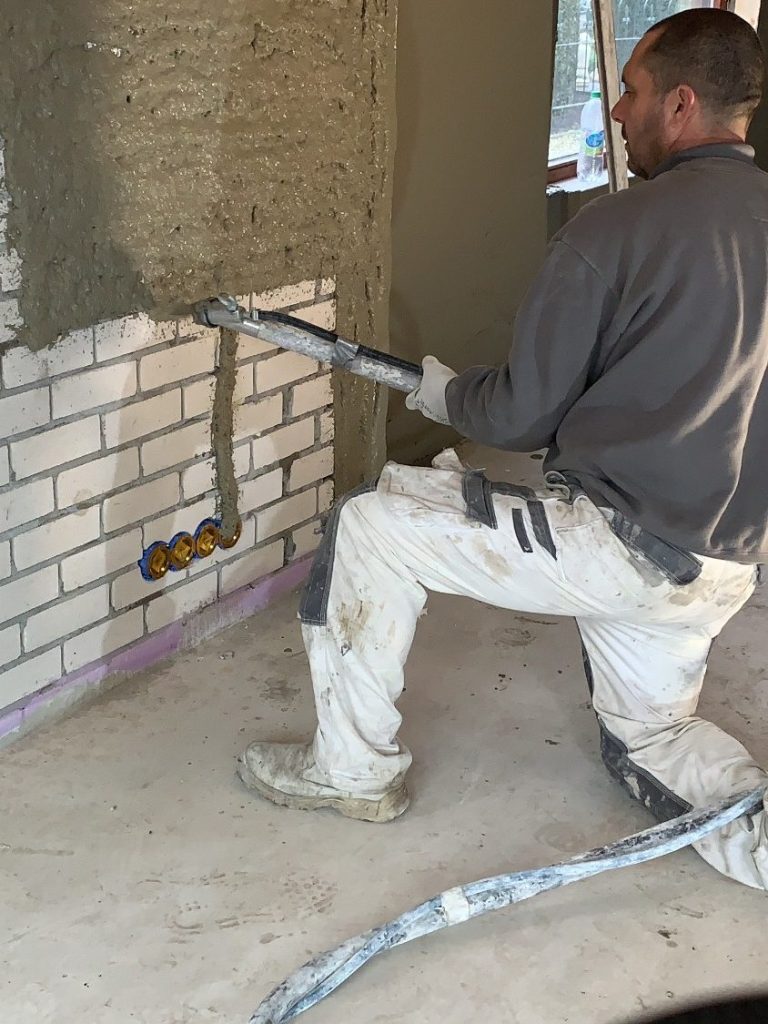 Een persoon brengt cement aan op een wand met een ritmo stucpomp, in een bouwomgeving.